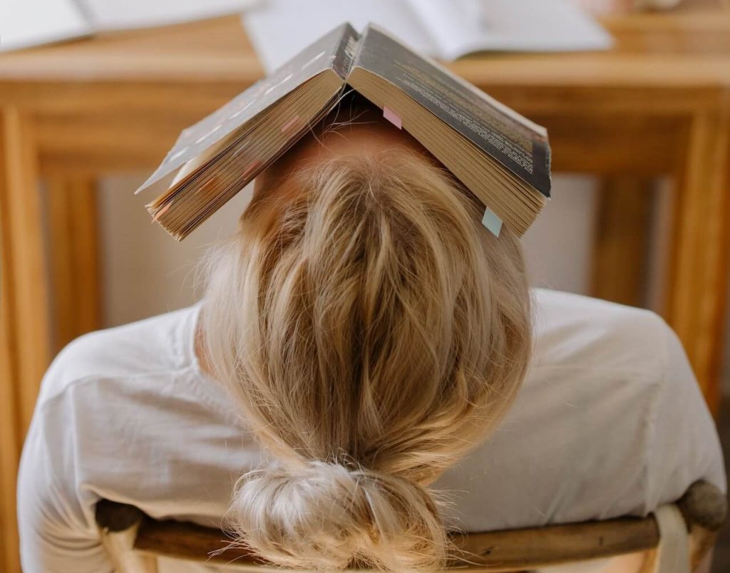 L'immagine di una donna stressata seduta su una sedia, con la testa inclinata all'indietro e un libro aperto appoggiato sul viso, raffigura la sensazione di stress.